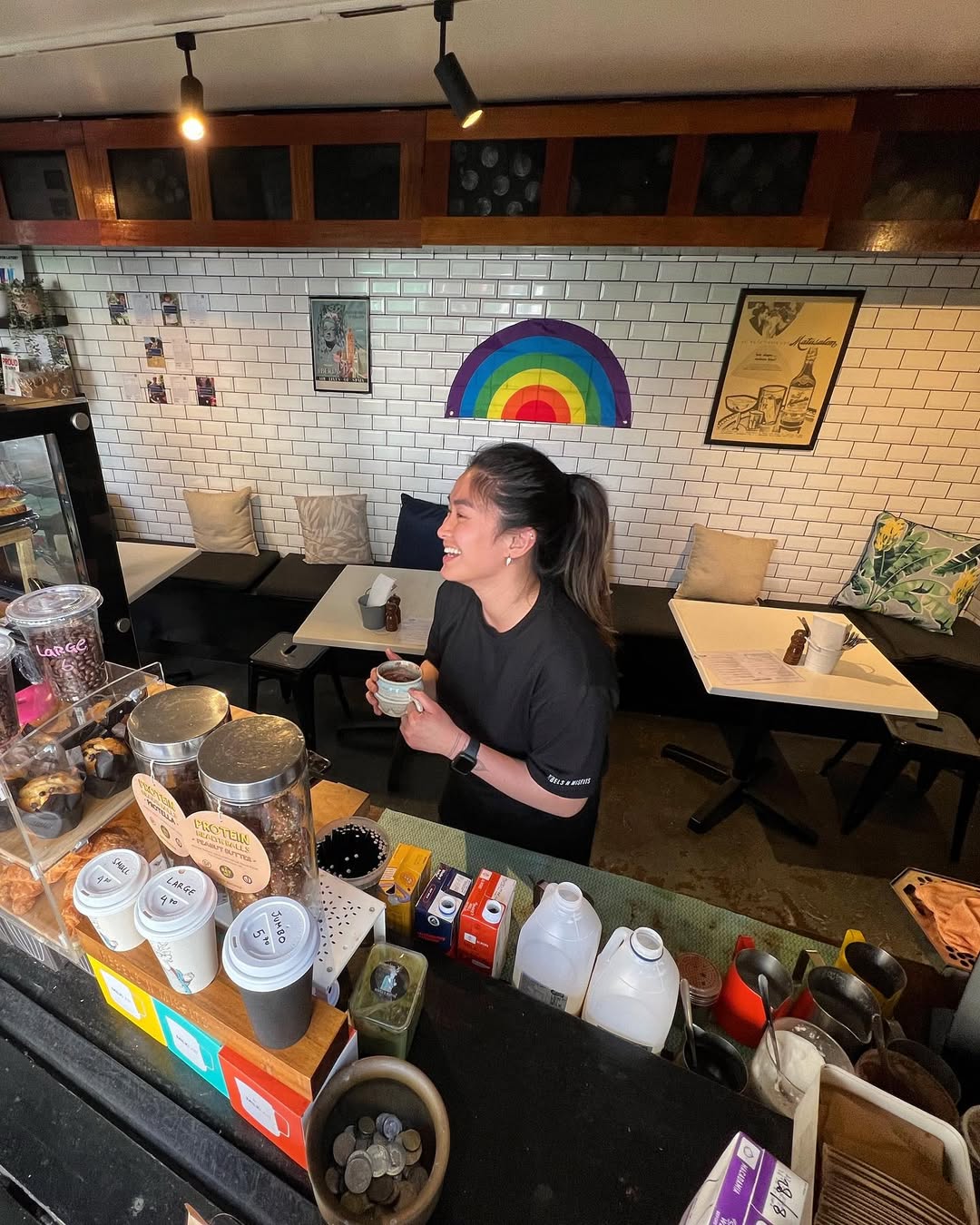 A smiling Rebels N Misfits barista with a coffee, behind the counter